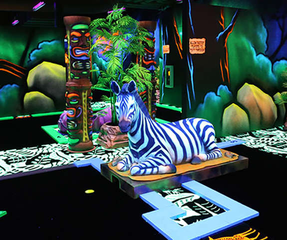 An overview of our blacklight mini golf course with a sitting zebra sculpture and African landscape..